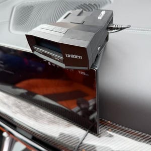 May include: A black Uniden radar detector mounted on a vehicle dashboard. The device features a rectangular housing with a front-facing display screen, control buttons on the top surface, and a power cable connected to the side of the unit.