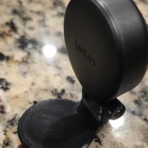 Puede incluir: Un soporte negro, circular y ajustable que cuenta con una base plana y redonda y un componente de disco vertical pivotante. El disco tiene la palabra VAVA grabada en su superficie. Todo el objeto está hecho de un material mate y texturizado.