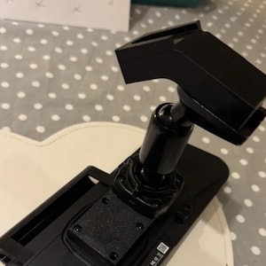 May include: A black plastic mounting bracket featuring a rectangular base with a central ball-and-socket joint connected to an angled, hollow rectangular head. The base includes a small square label with printed text and a visible FCC logo.