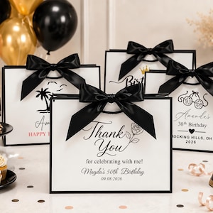 Può includere: Sacchetti regalo rettangolari bianchi con bordi neri e grandi fiocchi in nastro di raso nero. La parte anteriore dei sacchetti presenta testi e grafiche stampate in nero, incluse frasi come Thank You for celebrating with me, Amanda's 30th Birthday e Magda's 50th Birthday con date.