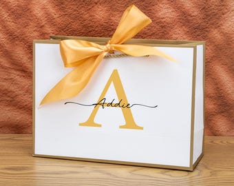 May include: White gift bag with gold trim, a gold satin bow, and the name "Addie" in a script font below a gold letter "A". The bag is sitting on a wooden surface.