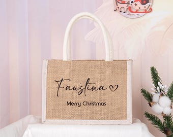 May include: A rectangular burlap tote bag with white trim and handles. The text "Faustina ♡ Merry Christmas" is printed in black on the front of the bag. The bag is sitting on a white surface.