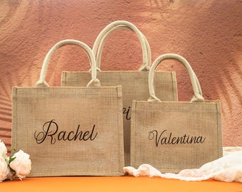 May include: Three brown burlap tote bags with cream colored handles. The bags are personalized with the names "Rachel", "Valentina", and "[unclear name]" in black cursive lettering.