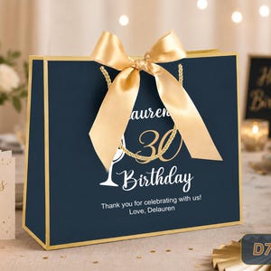 May include: A navy blue gift bag with gold trim and a large gold satin ribbon bow. The front features the text "Lauren's 30 Birthday" in white and gold, a white wine glass graphic, and the text "Thank you for celebrating with us! Love, Delauren".