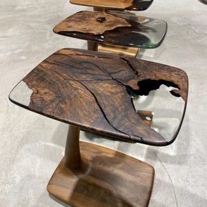May include: A series of side tables featuring live-edge dark brown wood slabs with natural cracks and voids filled with clear resin. Each table has a smooth, rounded rectangular top supported by a single solid wood pedestal and a flat base.