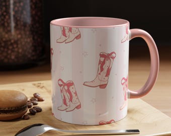 May include: Pink and white ceramic mug with a pattern of pink cowboy boots and stars. The mug has a pink handle.