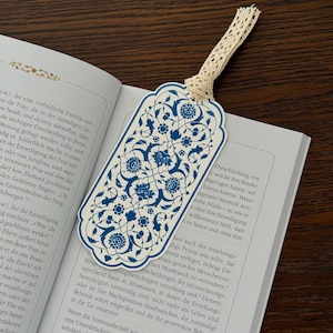 Könnte beinhalten: Ein Lesezeichen mit blauem Blumenmuster auf cremefarbenem Hintergrund. Oben ist ein cremefarbenes Spitzenband befestigt. Das Lesezeichen liegt in einem geöffneten Buch.
