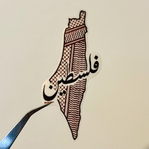 Könnte beinhalten: Ein Aufkleber in Form von Palästina, mit einem traditionellen Keffiyeh-Muster in Burgunderrot und Weiß. Das arabische Wort "فلسطين" ist in schwarzer Schrift geschrieben. Der Aufkleber wird von einer Pinzette gehalten.