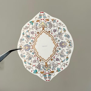 Könnte beinhalten: Ein weißes, ovales dekoratives Keramikstück mit einem floralen Design in Blau, Lila und Gold. Die Mitte zeigt ein goldumrandetes, rautenförmiges Design. Das Stück wird von einer Pinzette vor grauem Hintergrund gehalten.
