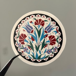 Könnte beinhalten: Ein runder Aufkleber mit floralem Design. Er zeigt eine zentrale Anordnung aus roten, blauen und grünen Blumen und Blättern, umgeben von einem schwarz-weißen Rand. Eine Pinzette hält den Aufkleber.