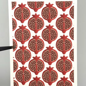 Könnte beinhalten: Eine weiße Karte mit einem sich wiederholenden Muster aus roten Granatapfel-Illustrationen. Jeder Granatapfel ist halbiert und zeigt die Samen. Das Design hat eine Vintage-Ästhetik. Der Künstlername, @suneylast, befindet sich unten rechts.