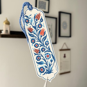 May include: A decorative bookmark with a blue braided cord. The bookmark features a floral design with red tulips and blue flowers on a white background. The bookmark has a unique shape and is held up by a pair of scissors.