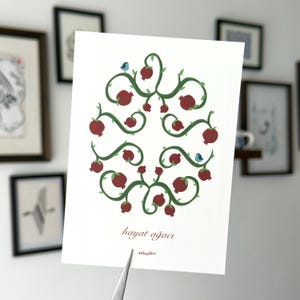 May include: A white card featuring a watercolor design of a pomegranate tree. The design includes red pomegranates, green vines, and two blue birds. The text "hayat ağacı" is written in a script font.