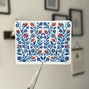 Könnte beinhalten: Ein rechteckiger Aufkleber mit floralem Design. Das Muster zeigt blaue und rote Blumen und Blätter auf weißem Hintergrund. Der Aufkleber wird von einer Pinzette gehalten.