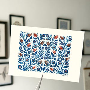 Pode incluir: Impressão de arte retangular com um padrão floral em tons de azul, vermelho e castanho. O design apresenta flores e folhas estilizadas em um arranjo simétrico. A impressão é segurada por uma pinça. Assinatura do artista visível.