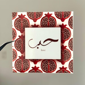 Könnte beinhalten: Quadratische dekorative Fliese mit einem Granatapfelmuster in Rot und Braun auf weißem Hintergrund. Ein kleineres weißes Quadrat in der Mitte zeigt arabische Kalligraphie und das Wort "love".