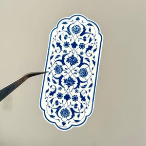 Könnte beinhalten: Ein weißer Aufkleber mit blauem Blumenmuster, gehalten von einer Pinzette. Der Aufkleber hat eine rechteckige Form mit gewölbter Ober- und Unterseite. Das florale Muster ist detailliert.