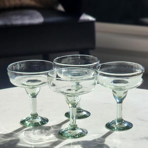 Pu&ograve; includere: Quattro bicchieri da margarita in vetro trasparente con bordi verdi. I bicchieri sono su una superficie bianca.