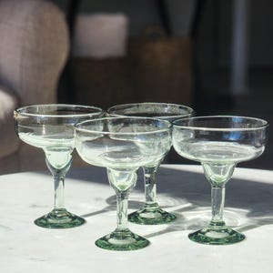 Pu&ograve; includere: Quattro bicchieri da margarita in vetro trasparente con bordi e basi verdi. I bicchieri sono disposti a semicerchio su una superficie bianca.