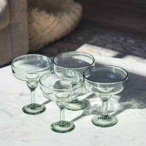 Pu&ograve; includere: Quattro bicchieri da margarita in vetro trasparente con bordi verdi sono disposti su una superficie di marmo bianco. I bicchieri sono vuoti e hanno una leggera sfumatura verde.