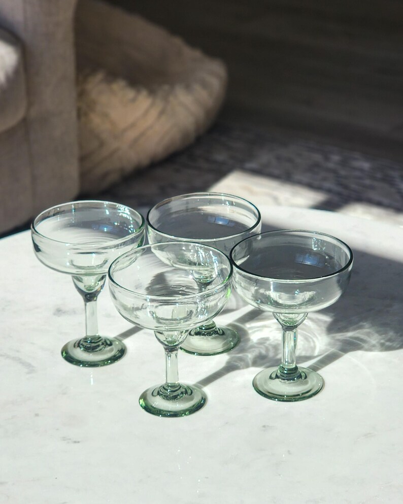 Pu&ograve; includere: Quattro bicchieri da margarita in vetro trasparente con bordi verdi sono disposti su una superficie di marmo bianco. I bicchieri sono vuoti e hanno una leggera sfumatura verde.