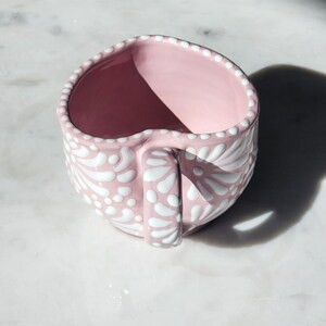 May include: A pink ceramic mug with a white floral design. The mug has a handle and is decorated with white dots.