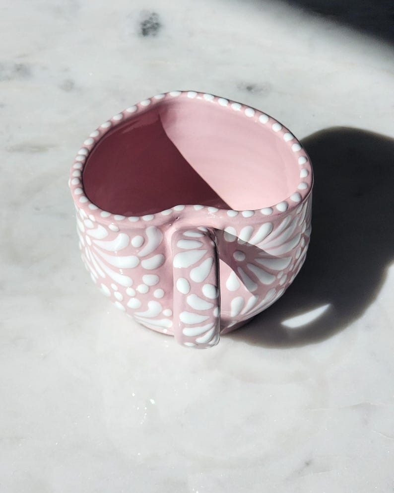 May include: A pink ceramic mug with a white floral design. The mug has a handle and is decorated with white dots.