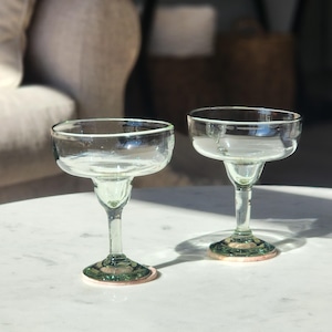 Pu&ograve; includere: Due bicchieri da margarita in vetro trasparente con bordi verdi e basi in rame. I bicchieri sono su una superficie bianca.