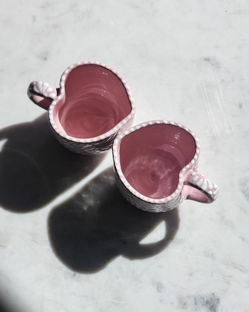 May include: Two pink heart-shaped mugs with white floral designs. The mugs are empty and sitting on a white marble surface.