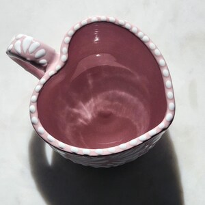 May include: A pink heart-shaped ceramic mug with a white polka dot design around the rim and handle.