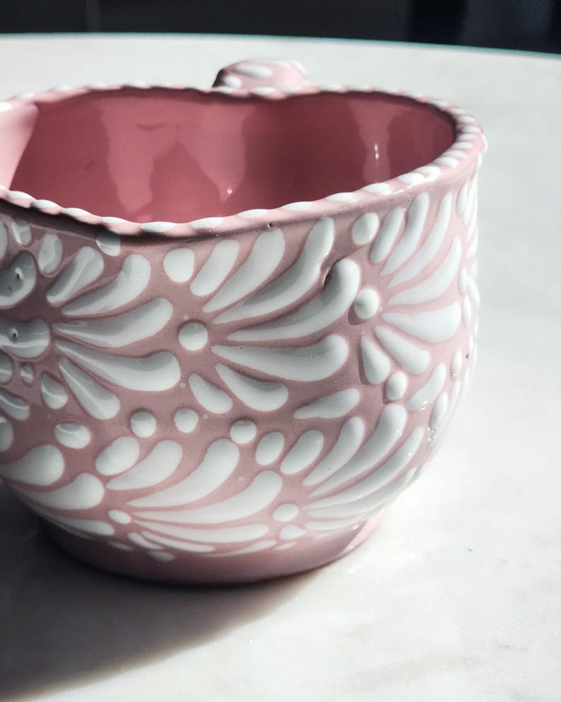 May include: A pink ceramic mug with a white floral design. The mug has a rounded shape and a handle.
