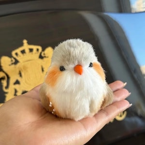 May include: A plush bird toy with a fluffy white chest, gray head, and orange accents on the cheeks and beak. The bird has small black eyes and is held in a hand. The toy is a cute, decorative item.