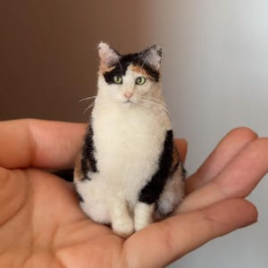 Könnte beinhalten: Eine Miniatur-Calico-Katzenfigur mit weißen, schwarzen und orangefarbenen Markierungen. Die Katze hat grüne Augen und sitzt aufrecht. Die Figur wird in einer Hand gehalten, was ihre geringe Größe und detaillierte Verarbeitung zeigt.
