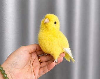 Puede incluir: Un pájaro amarillo brillante con la cola con punta blanca está posado en una mano. El pájaro tiene un pico pequeño y de color claro y un ojo negro. El pájaro está hecho de un material suave y esponjoso, sobre un fondo gris claro.