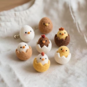 Op de afbeelding: Een verzameling handgemaakte vilten kuikens en eieren in verschillende kleuren. De kuikens zijn geel, bruin en wit, sommige komen uit gebarsten eierschalen. De eieren en kuikens zijn gerangschikt op een wit oppervlak.