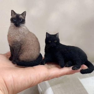 Op de afbeelding: Twee miniatuur kattenbeeldjes van naaldvilt. De ene is een Siamees met een lichtbruin lichaam, donkerbruin gezicht en blauwe ogen, zittend. De andere is een effen zwarte kat met gele ogen, die ontspannen en uitgestrekt op de buik ligt.