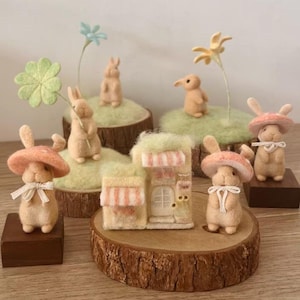 Peut inclure: Une collection de figurines en laine feutrée représentant des lapins avec des chapeaux en forme de champignon, une petite maison et des fleurs. Les lapins sont beiges avec des chapeaux roses et des nœuds blancs. La maison a un auvent rayé.