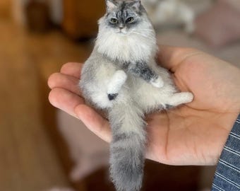 May include: A miniature, fluffy cat figurine with gray and white fur. The cat is posed sitting with its legs extended, and its long tail is visible. The cat's fur is detailed, and it has green eyes. The figurine is held in a person's hand.