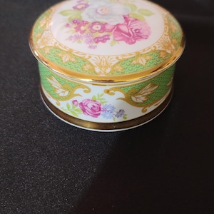 May include: A round porcelain trinket box with a floral design. The lid features a green and gold border with a white background and pink, purple, and white flowers. The bottom of the box has a similar design with a green and gold border and a white background with pink, purple, and white flowers.