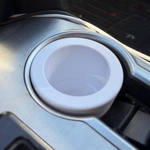 May include: White plastic cup holder in a car. The cup holder is in a silver metal console.