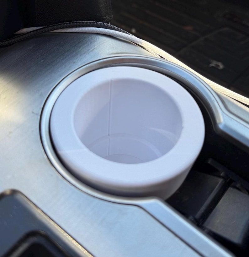 May include: White plastic cup holder in a car. The cup holder is in a silver metal console.