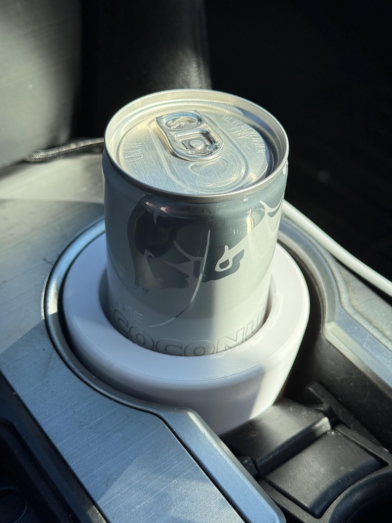 May include: A silver can of Red Bull Coconut Edition in a white cup holder in a car.