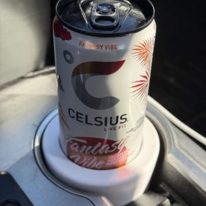May include: A white can of Celsius energy drink with the flavor "Fantasy Vibe" printed on the can. The can is in a white cup holder in a car.