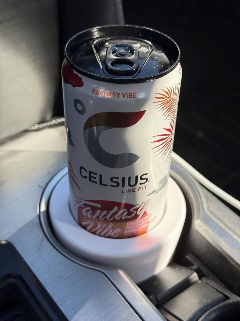 May include: A white can of Celsius energy drink with the flavor "Fantasy Vibe" printed on the can. The can is in a white cup holder in a car.