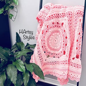 May include: A light pink crochet blanket with a central circular pattern and a scalloped edge, draped over a black ladder. The text "HArtsy Styles" is visible on the wall. The blanket is handmade.
