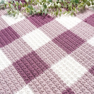 May include: A handmade crochet blanket with a checkered pattern in shades of purple, white, and light purple. The blanket features a textured stitch and is adorned with the text "HArtsy Styles".