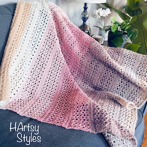 May include: A crocheted blanket featuring a gradient of colours including beige, white, pink, and mauve. The textile has a repeating openwork stitch pattern with vertical textured stripes. The text HArtsy Styles is printed in white on the lower left.
