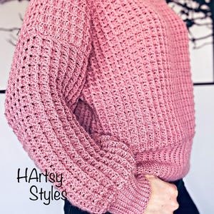 May include: A pink crocheted sweater with a waffle stitch pattern. The sweater has long sleeves and a relaxed fit. The text "HArtsy Styles" is visible on the sweater.