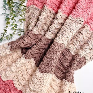 Op de afbeelding: Een handgemaakte gehaakte deken in tinten roze, crème en bruin. De deken heeft een gestructureerd, golvend patroon. De tekst "HArtsy Styles" is zichtbaar in de rechter benedenhoek. Deze gezellige deken is perfect voor de interieurdecoratie.