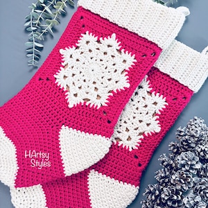 以下が含まれることがあります： 2つの手作りのクリスマスストッキング。ストッキングは明るいピンク色で、雪の結晶のデザインとカフスなど、白いアクセントが付いています。「HArtsy Styles」の文字がストッキングに表示されています。ストッキングはかぎ針編みで作られています。